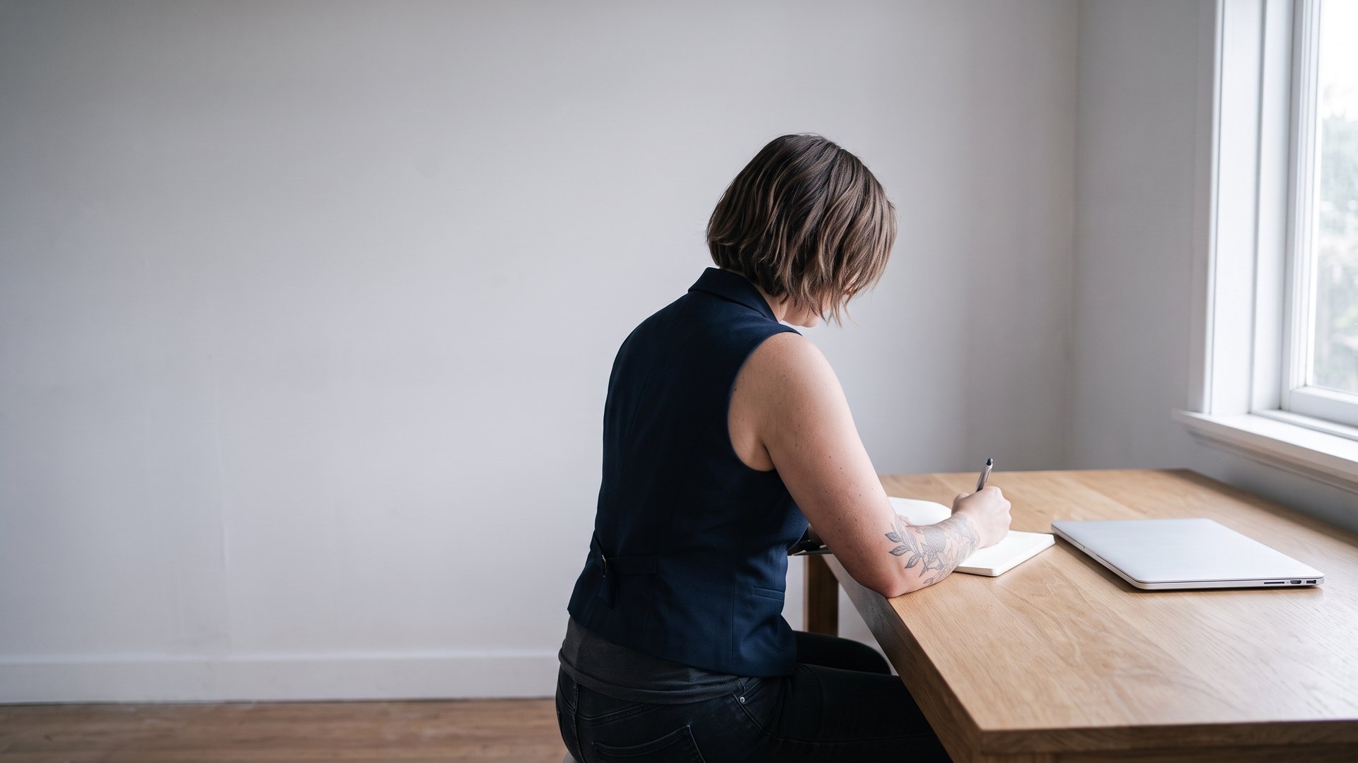 Emma Hanover seated at a plain oak desk beside a window, writing in a notebook with a closed laptop next to her